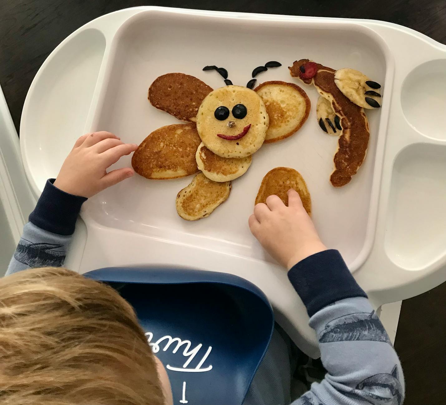 Child playing with a butterfly-shaped pancake breakfast on a highchair tray, creating a fun and artistic meal time.