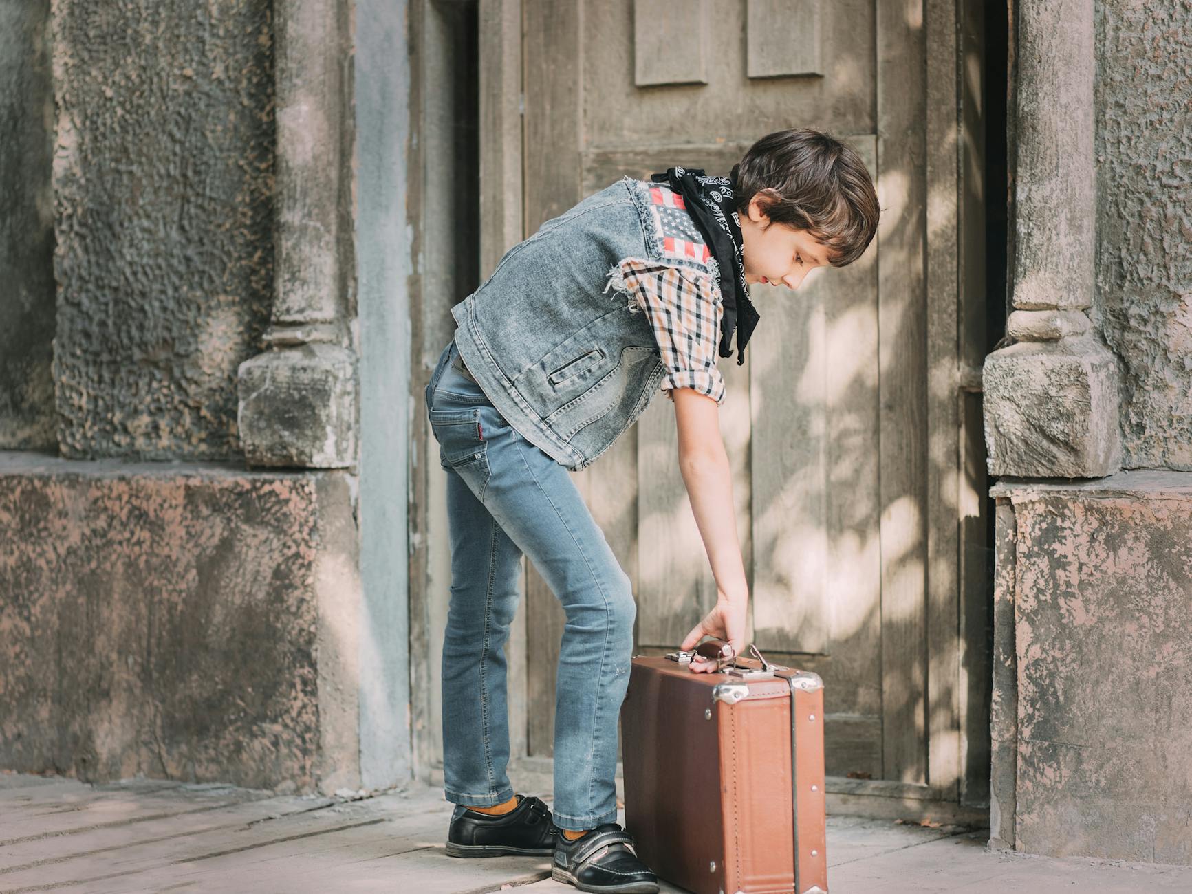 A child dressed in casual fashion holding a vintage suitcase outside an old building.