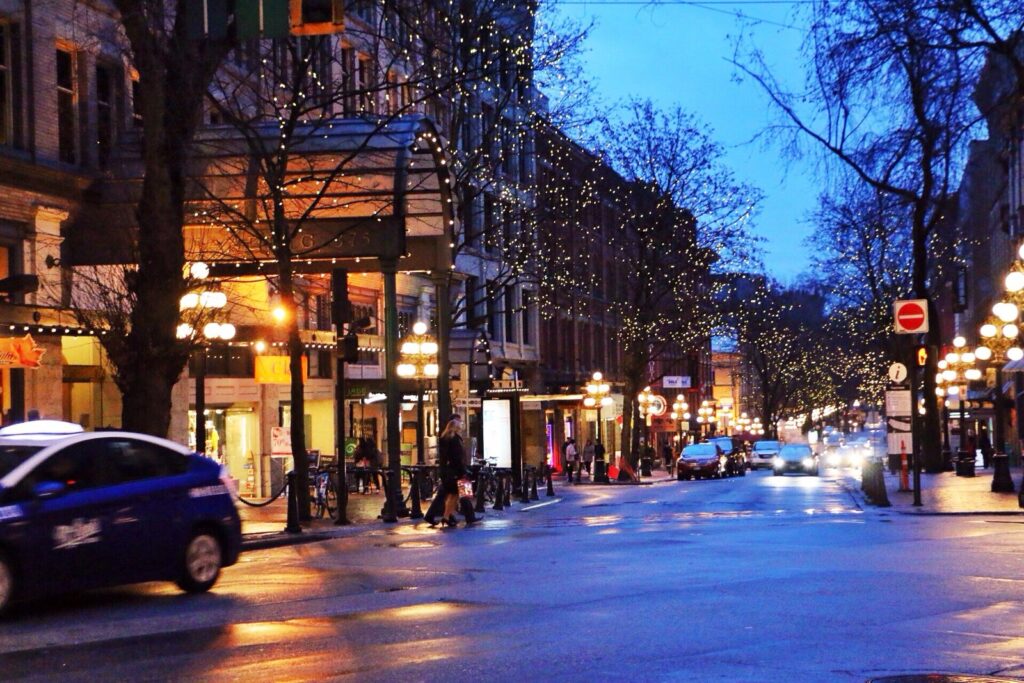 Water Street in Gastown at dusk with lit shopfronts, street lamps and passing cars