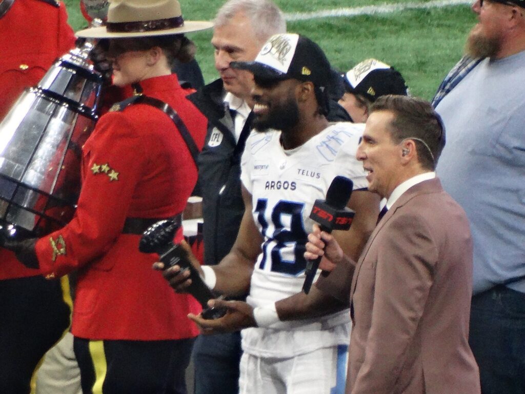 Toronto Argonauts player holding the Grey Cup trophy beside an RCMP officer in red serge on the BC Place field