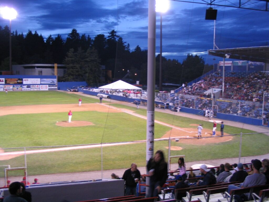 Nat Bailey Stadium at dusk during a Vancouver Canadians game, with the lights on and tall trees behind the outfield