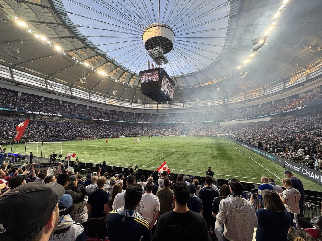 Packed BC Place crowd with flags under the centre-hung scoreboard during a Whitecaps night match